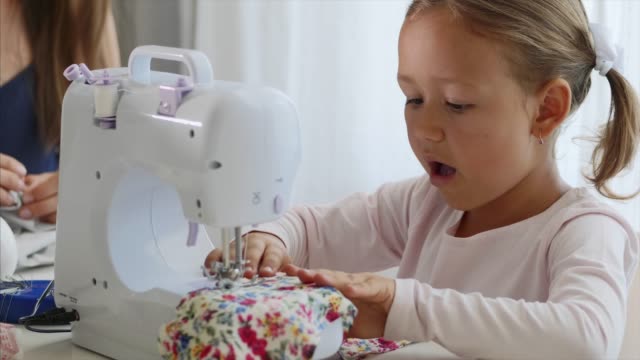 A-little-girl-is-sewing-fabric-and-talking.-Mom-is-at-the-background.-Close-up.