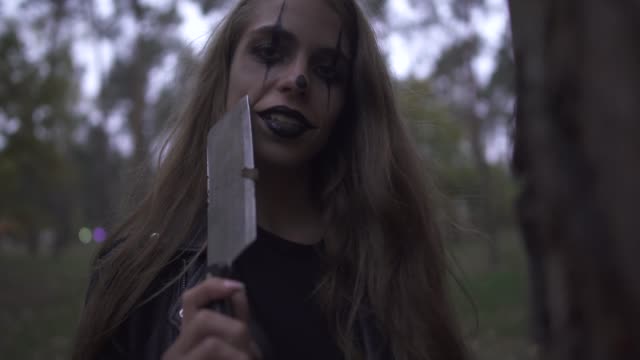 Crazy-young-woman-with-Halloween-clown-makeup-on-her-face-holding-big-knife-in-hand,-laughing.
