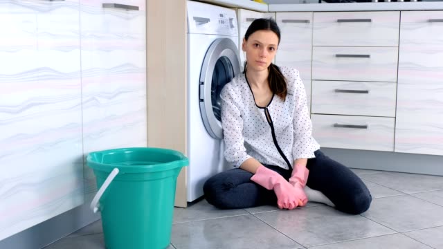 Mujer-cansada-con-guantes-de-goma-rosa-en-el-suelo-de-la-cocina-después-de-limpiar-mira-la-cámara.
