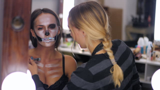 Makeup-artist-paints-greasepaint-for-Halloween-in-studio.Woman-drawing-a-glamorous-skull-with-rhinestones-and-sequins-on-a-beautiful-young-girl-with-long-hair.Reflection-in-the-Mirror.Slow-motion