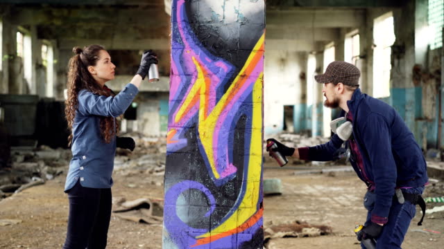 Zwei-erfahrene-Graffiti-Künstler-bärtigen-Kerl-und-attraktive-junge-Frau-arbeiten-zusammen-in-verlassenen-Lagerhaus-Dekoration-alte-Spalte-mit-abstrakten-Bild.