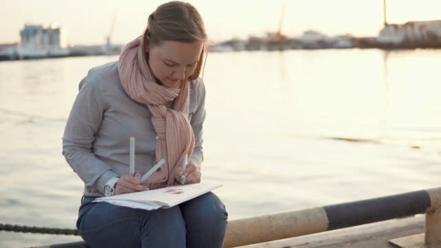 Young-woman-is-sitting-near-pier-in-spring-evening-and-drawing-by-pencils