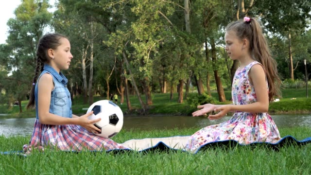 Zwei-kleine-Mädchen-spielen-mit-einem-Fußball-auf-dem-Rasen-sitzen.-Sie-sitzen-bei-Sonnenuntergang-durch-den-Park-gegenüber-einander-und-werfen-den-Ball-zu-einander.