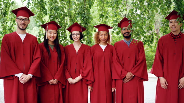 Retrato-de-grupo-multiétnico-de-los-estudiantes-graduados-de-pie-al-aire-libre-llevando-vestidos-de-rojo-y-placas-de-mortero,-sonriendo-y-mirando-a-cámara.