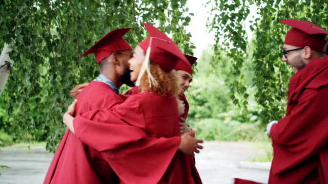 Excited-young-people-graduating-students-in-gowns-and-hats-are-hugging-congratulating-each-other-on-graduation,-laughing-and-celebrating-end-of-academic-year.