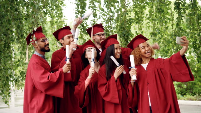 Muy-contentos-los-estudiantes-graduados-están-tomando-selfie-con-smartphone,-jóvenes-están-agitando-diplomas,-posando,-sonriendo-y-riendo.-Concepto-de-educación-y-el-éxito.