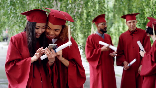 Atractivas-mujeres-jóvenes-graduados-felizes-viendo-fotos-en-el-teléfono-inteligente-y-hablar-en-el-día-de-la-graduación-mientras-sus-compañeros-están-chateando-en-fondo.