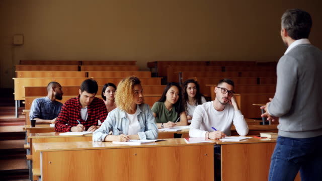 Tutor-is-talking-to-students-sitting-at-desks-in-spacious-college-lecture-hall,-young-people-in-casual-clothes-are-asking-questions.-Education-and-university-concept.