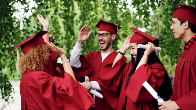 Multiracial-group-of-young-people-students-are-doing-high-five-on-graduation-day-wearing-traditional-clothes-hats-and-gowns.-Millennials,-youth-and-education-concept.