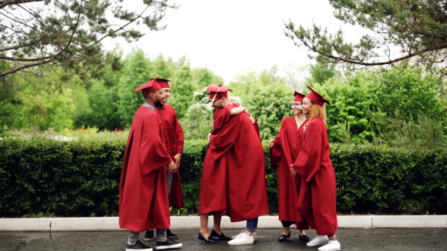 Multiethnic-group-of-friends-graduates-are-hugging,-doing-high-five-and-dancing-in-mortar-boards-and-gowns-traditional-graduation-clothes.-Youth-and-fun-concept.