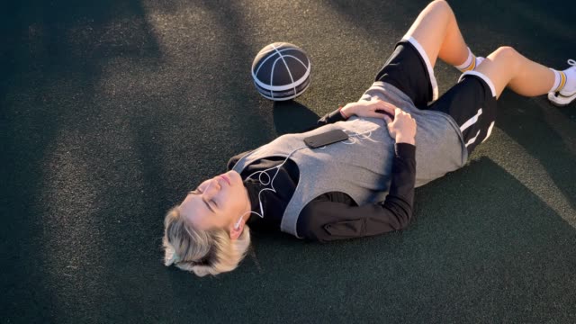 Beautiful-blonde-female-basketball-player-lying-on-court-and-listening-music,-woman-relaxing-in-open-air-during-sunset