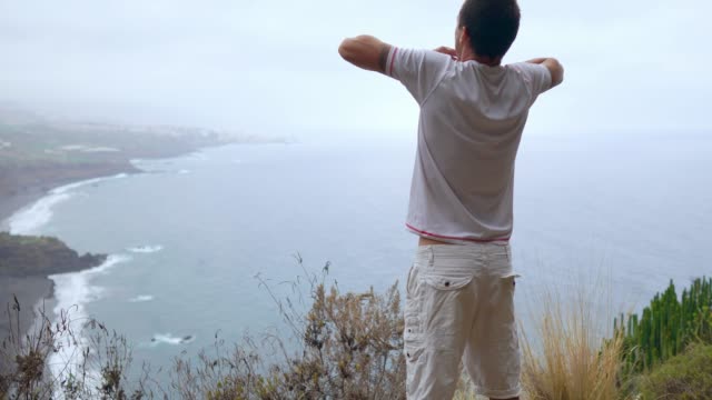 Ein-Mann-steht-am-Rand-einer-Klippe-mit-Blick-auf-den-Ozean-erhebt-seine-Hände-und-atmet-die-Luft-während-yoga