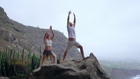 A-man-and-a-woman-standing-on-the-edge-of-a-cliff-overlooking-the-ocean-raise-their-hands-up-and-inhale-the-sea-air-during-yoga