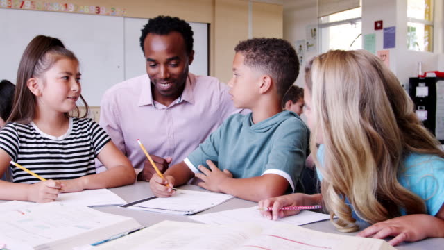 Male-elementary-school-teacher-comes-to-kidsÍ-desk-to-help