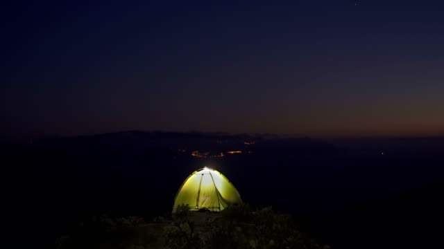 the-tourist-tent-glows-in-the-night-on-the-high-coast-of-the-ocean-opposite-the-night-lights-of-the-other-island.-Strong-gusts-of-wind-rocking-the-tent