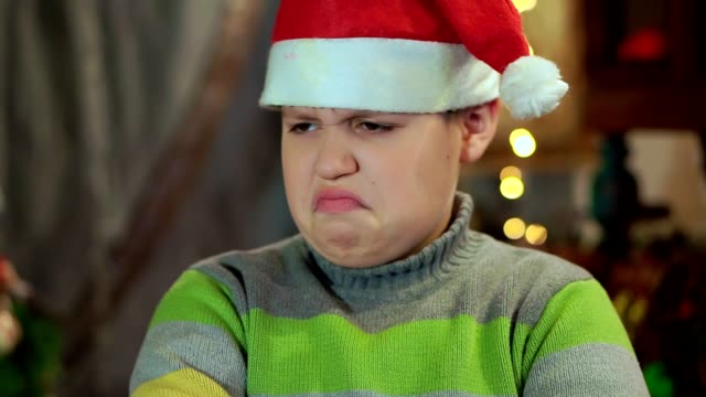 The-boy-in-the-sweater-and-the-hat-of-Santa-Claus-is-upset-and-offended.-He-has-no-gift.-Close-up,-against-the-background-of-Christmas-lights