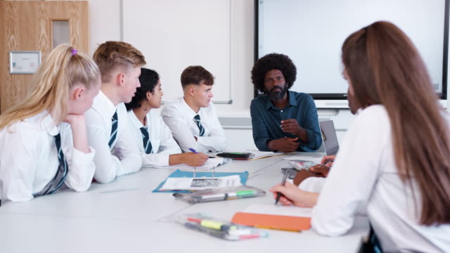 Männlichen-High-School-Lehrer-mit-Teenager-Schüler-tragen-einheitliche-Lehre-Lektion-am-Tisch-sitzen