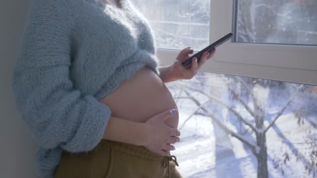moderne-Elternschaft,-Mann-Hände-streicheln-Bauch-der-schwangeren-Frau-mit-Handy-in-Nahaufnahme-Arm-in-natürliches-Licht-in-der-Nähe-von-Fenster