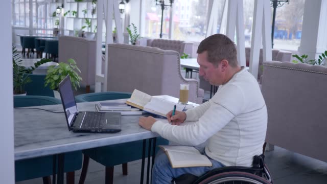 Fernstudium-der-ungültig,-erfolgreiche-kranke-Schüler-männlich-auf-Rollstuhl-arbeiten-mit-Laptop-von-Online-Lektionen-zu-lernen-und-sich-Notizen-macht,-in-notebook