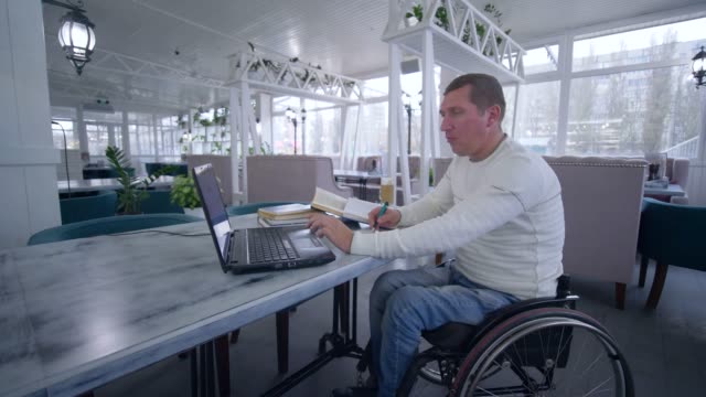 Educación-en-línea,-hombre-de-estudiantes-con-discapacidad-en-silla-de-ruedas-utiliza-tecnología-portátil-moderno-para-aprender-de-lecciones-en-línea-y-libros-haciendo-notas-en-sesión-primer-cuaderno-en-la-mesa-de-café
