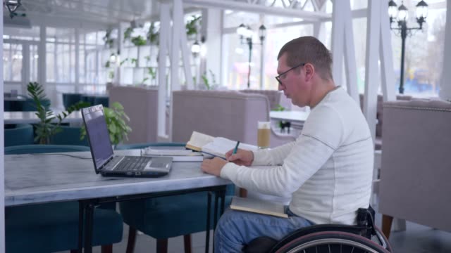 online-teaching,-aching-man-on-wheelchair-is-learning-for-self-development-uses-modern-technology-for-distance-education-and-makes-notes-in-notebook
