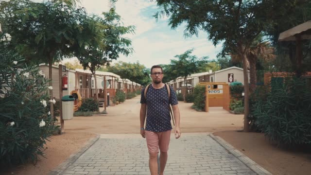 Bearded-tourist-man-is-walking-on-yard-of-resort-area-with-small-summer-cottages