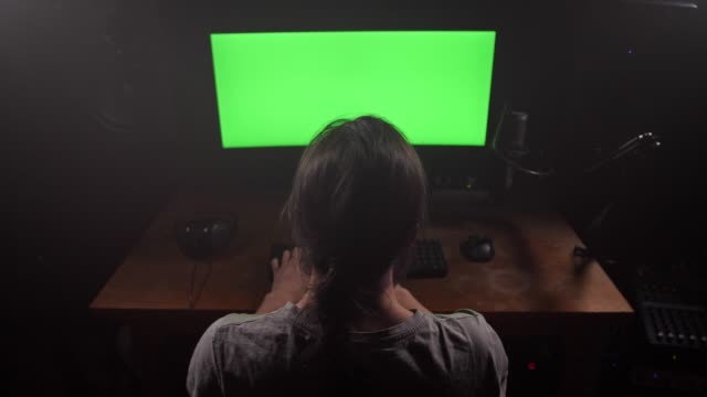 Ein-Mann-mit-langen-Haaren-druckt-Text-auf-seiner-Tastatur,-er-hat-einen-grünen-Bildschirm-auf-seinem-Monitor.-4K-Slow-Mo