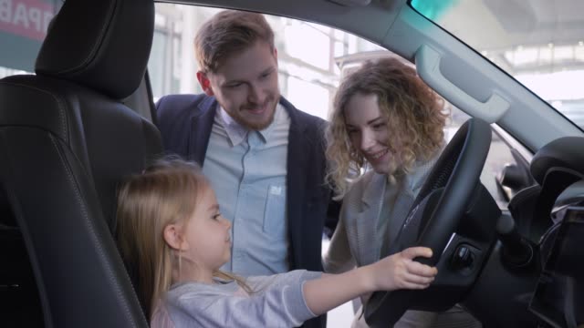 Lächeln-kleine-Mädchen-hinter-dem-Steuer-des-neuen-Fahrzeugs-zusammen-mit-Mama-und-Papa-beim-Kauf-von-Familienauto-beim-Händler