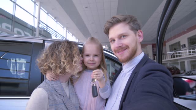 consumer-family-with-cute-little-daughter-with-keys-take-pictures-phone-on-telephone-near-new-purchased-machine-in-car-showroom
