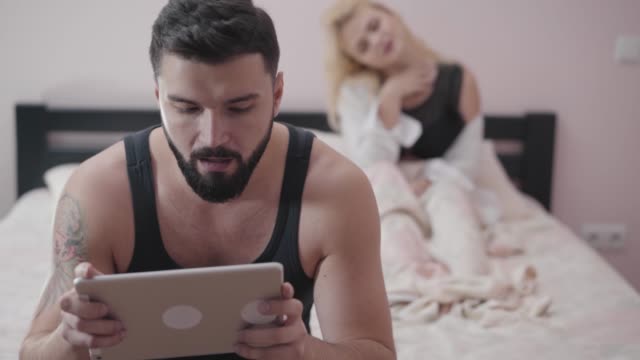 Portrait-of-adult-Caucasian-man-sitting-on-bed-and-playing-video-games-using-tablet.-Seductive-blond-woman-at-the-background-waiting-for-her-boyfriend-or-husband.-Games-addiction,-marriage-problems.