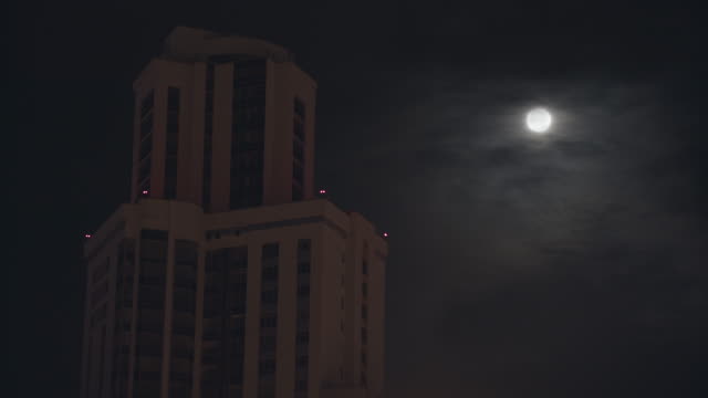 Night-cityscape-with-a-high-building-on-dark-sky-background.-Stock-footage.-Full-moon-on-dark-cloudy-sky