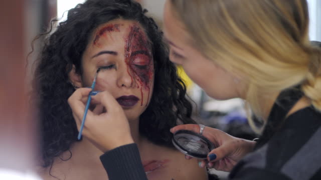 Make-up-artist-make-the-girl-halloween-make-upin-studio.Halloween-face-art.Woman-applies-on-professional-greasepaint-on-the-face-of-spanish-girl.War-paint-with-blood,-scars-and-wounds.Slow-motion