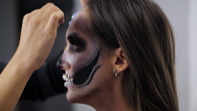 Makeup-artist-paints-greasepaint-for-Halloween-in-studio.-Woman-drawing-a-glamorous-skull-with-rhinestones-and-sequins-on-a-beautiful-young-girl-with-long-hair.-Slow-motion