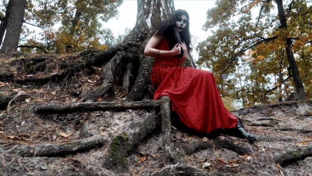 Woman-with-scary-halloween-make-up-in-red-dress-sitting-near-tree-in-the-forest-park-outdoors