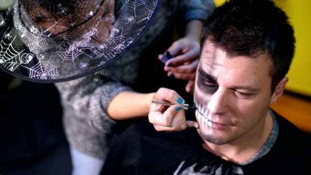 Halloween-Party,-close-up,-Make-up-Artist-zieht-eine-schreckliche-Make-up-auf-dem-Gesicht-eines-Mannes-für-eine-Halloween-Party.-im-Hintergrund-sieht-man-die-Landschaft-im-Stil-von-Halloween