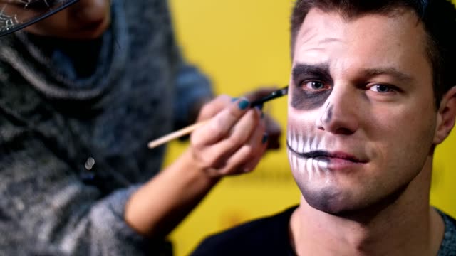 Halloween-party,-close-up,-make-up-artist-draws-a-terrible-makeup-on-the-face-of-a-man-for-a-Halloween-party.-in-the-background-the-scenery-in-the-style-of-Halloween-is-seen