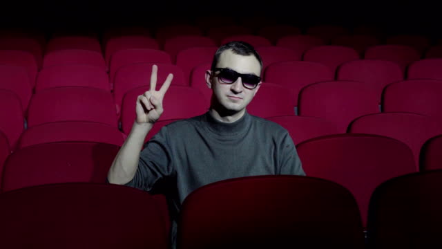 Single-man-sitting-in-comfortable-red-chairs-in-dark-cinema-theater-and-doing-victory-sign-with-fingers