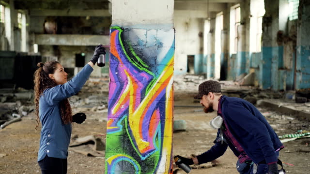 Kreatives-Team-von-zwei-städtischen-Maler-zeichnen-Graffiti-mit-Sprühfarbe-beim-alten-Industriehalle-mit-zerstörten-schmutzige-Wände-und-Fenster-dekorieren.