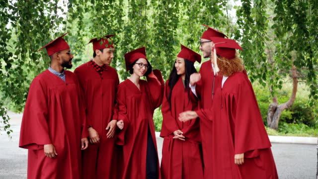 Retrato-de-estudiantes-graduados-emocionados-multiétnicos-grupo-permanente-al-aire-libre-en-vestidos-de-rojo-y-placas-de-mortero-y-hablando-y-mostrando-los-pulgares-para-arriba-y-mirando-a-cámara.