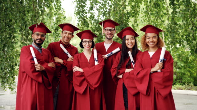 Portrait-of-group-of-graduating-students-holding-diplomas-and-standing-outdoors-with-arms-crossed-wearing-gowns-and-mortar-boards,-smiling-and-looking-at-camera.