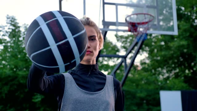 Schöne-blonde-Frau-zeigt-Basketball-in-Kamera,-Profi-Spieler-stehen-im-Park-tagsüber-Hoop-im-Hintergrund