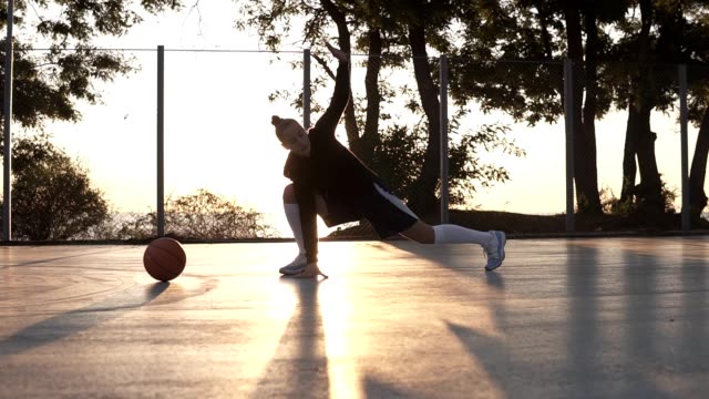 Caucasian-young-fit-woman-in-hoodie-and-shorts-warming-up-in-morning-on-basketball-court.-Stretching-her-legs-before-basketball-training.-Morning-dusk