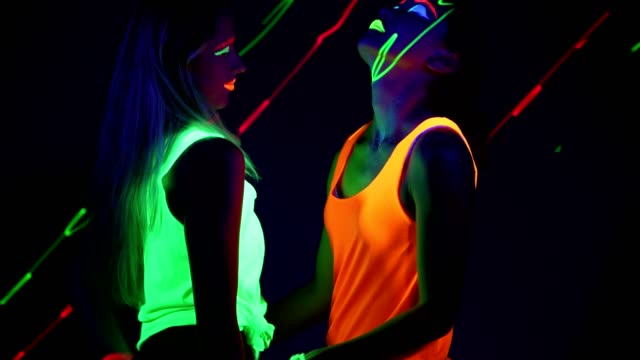 Women-with-UV-face-paint,-laser,-glowing-bracelets,-glowing-clothing-dancing-together-in-front-of-camera,-Half-body-shot.-Caucasian-and-asian-woman.-.
