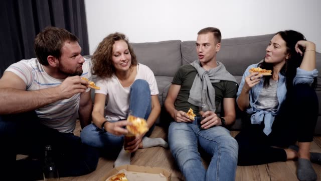Essen,-Freizeit-und-Glück-Konzept---vier-lächelnde-junge-Menschen-zu-Hause-Pizza-essen-und-trinken-ein-Bier-auf-dem-Boden-sitzend.-Vorderansicht