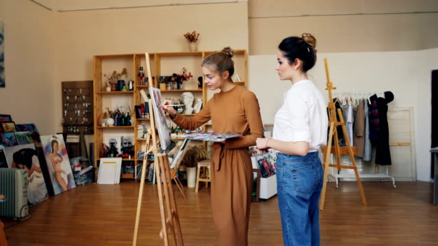 Slender-young-woman-art-student-is-painting-working-at-picture-and-talking-to-her-friendly-teacher-standing-indoors-in-modern-workshop.-Fine-arts-and-education-concept.