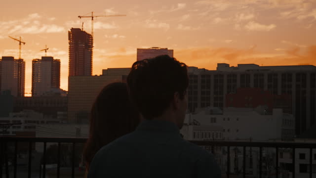 Rear-View-Of-Couple-On-Rooftop-Terrace-Looking-Out-Over-City-Skyline-At-Sunset