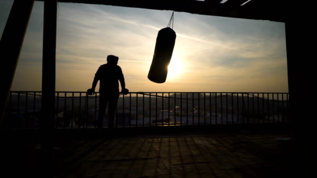 Boxeador-en-la-terraza-después-de-entrenamiento-mirando-la-ciudad,-bolso-de-balanceo,-hermosa-puesta-de-sol,-deportista-practicando-entrenamiento-power,-chico-fuerte-duro-ejercicio,-ejercicios-de-fuerza,-entrenamiento,-mano,-soleado-día.