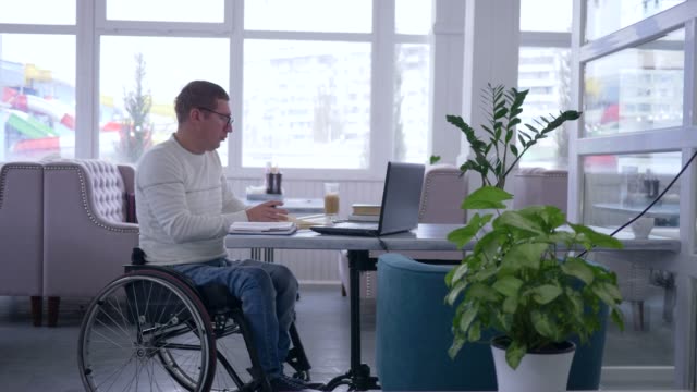 developing-of-Disabled-mature-man-in-wheelchair-into-glasses-works-on-laptop-during-distance-online-learning-sitting-at-table
