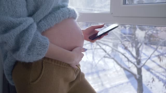 moderne-mobile-Technologie-für-Schwangere,-Mutterschaft-Weibchen-mit-großen-Bauch-mit-smart-Telefon-gegen-Fenster-im-Sonnenlicht-an-Wintertag