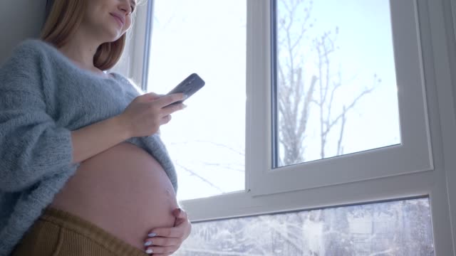 smiling-pregnant-female-with-big-tummy-uses-modern-mobile-technology-while-waiting-baby-against-window-in-sunlight-on-winter-day